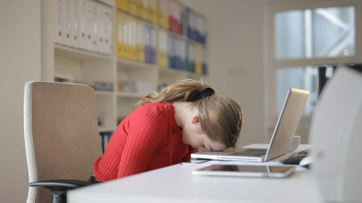 A tired woman in a red sweater leans her head on a desk with a laptop, symbolizing workplace fatigue.