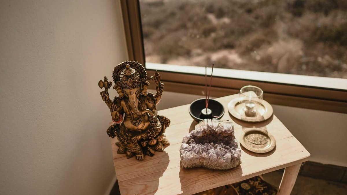 A Ganesha statue with incense sticks and an amethyst crystal on a wooden table by a window.