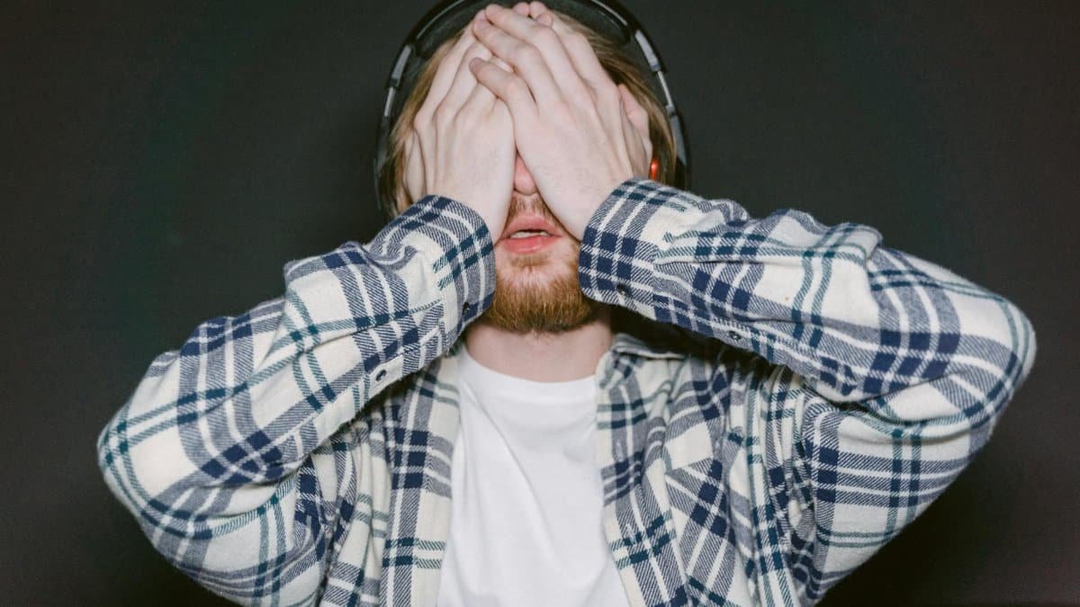 Young man wearing headphones and plaid shirt covering eyes, expressing stress or fatigue indoors.