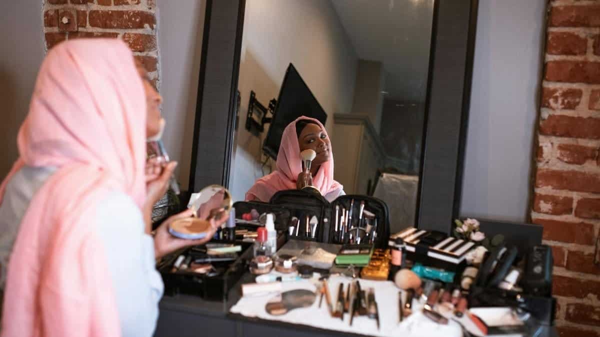 A woman in a headscarf applying makeup using a mirror, surrounded by cosmetic products.