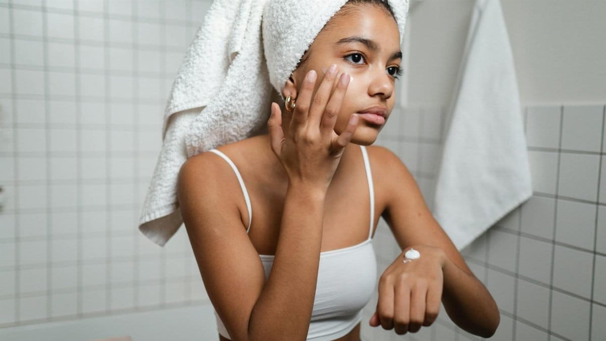 A young woman in a bathroom applying skincare cream with a towel on her head, focusing on self-care.