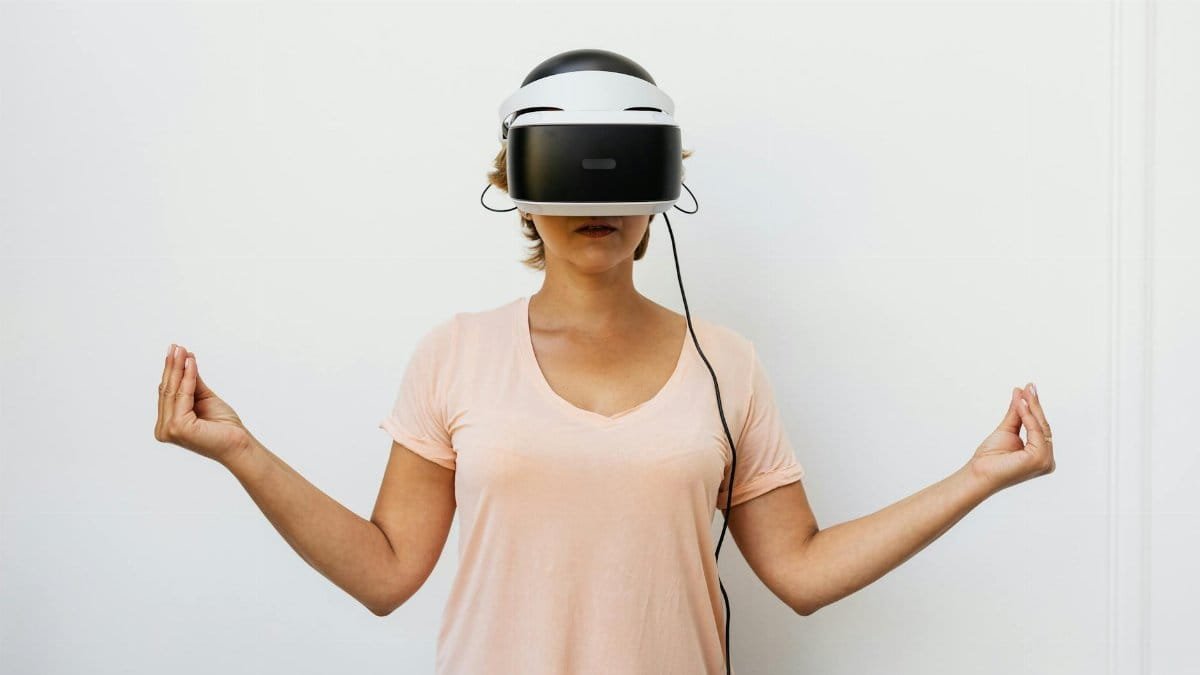 A woman wearing virtual reality goggles stands with a meditation gesture indoors.