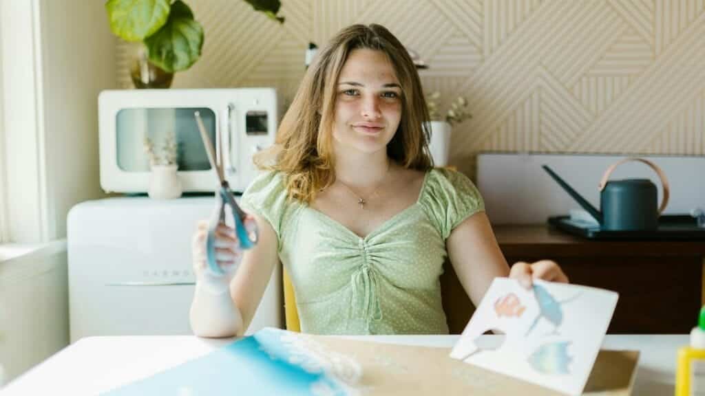 Teen girl crafting indoors, holding scissors, in a light-filled room, focused on creativity.
