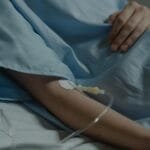 Close-up of a patient in a hospital bed with an IV drip attached to the arm.