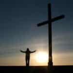 Silhouette of a person with open arms beside a cross at sunset, symbolizing faith and spirituality.