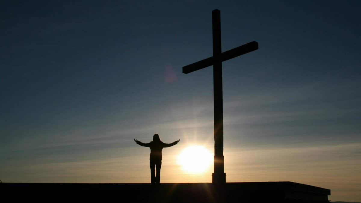 Silhouette of a person with open arms beside a cross at sunset, symbolizing faith and spirituality.