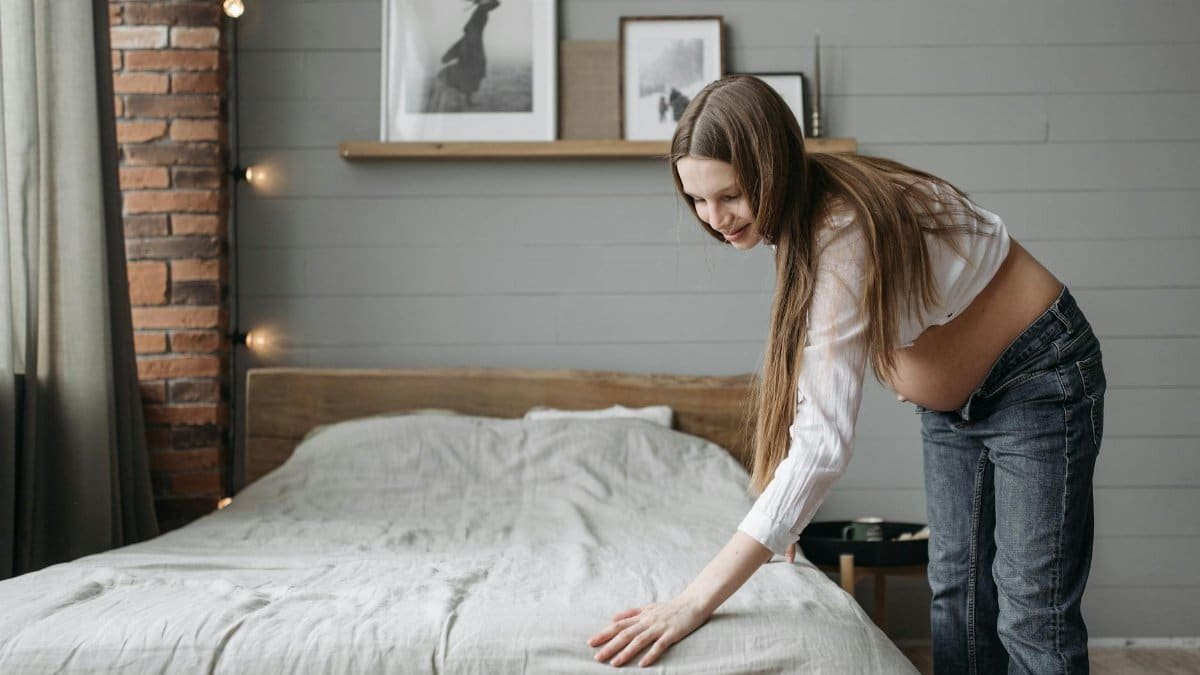 Expectant mother tidying a stylish, minimalist bedroom with framed art and soft lighting.