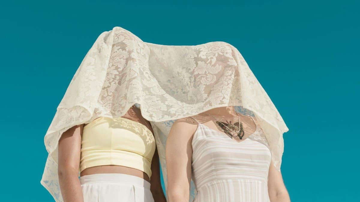 Minimalist shot of two women under a white lace veil against a clear blue sky.