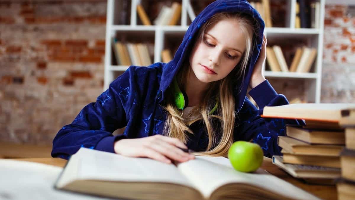 Teenage girl in blue hood reads in library, headphones and apple by her side.