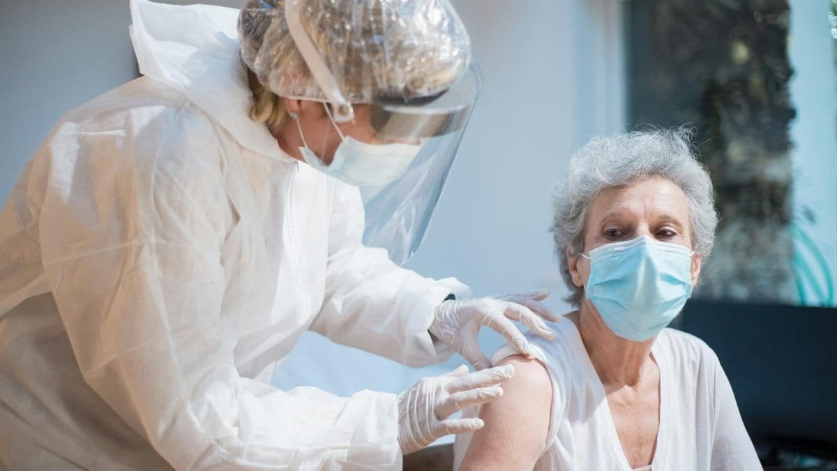 Nurse in protective gear giving vaccine to senior woman indoors.