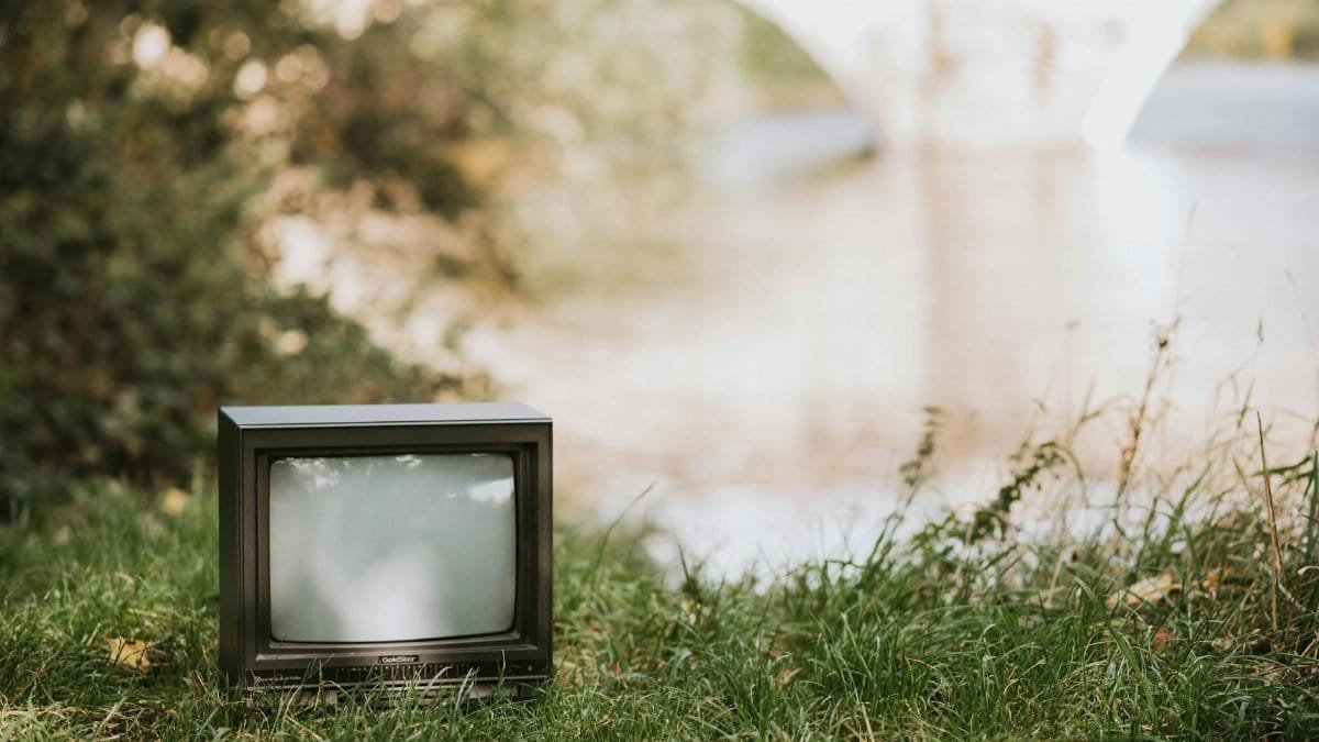 Small retro television placed on lakeside near calm water with green bushes and bridge on blurred background in sunny nature