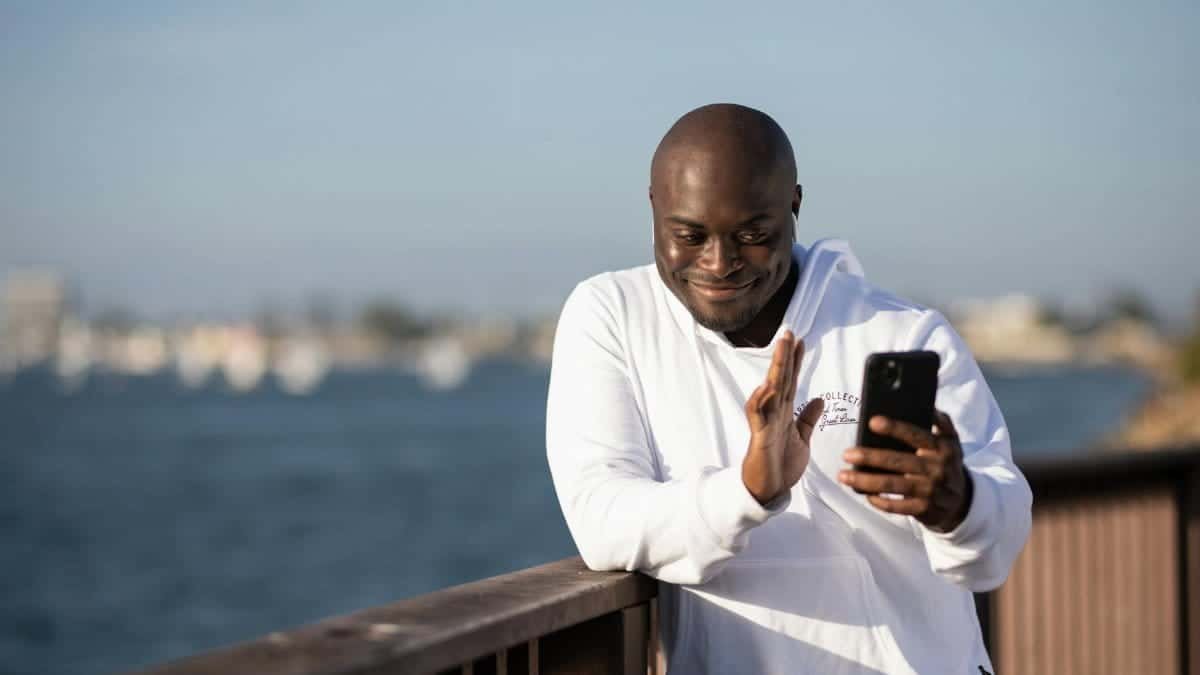 Bald man in white hoodie making a video call by the waterfront.