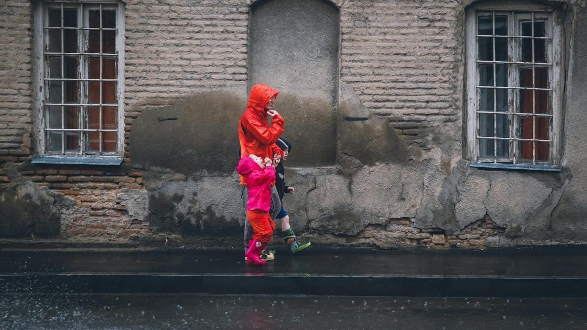 A family walks along a rainy street beside a historic building, wearing colorful raincoats.