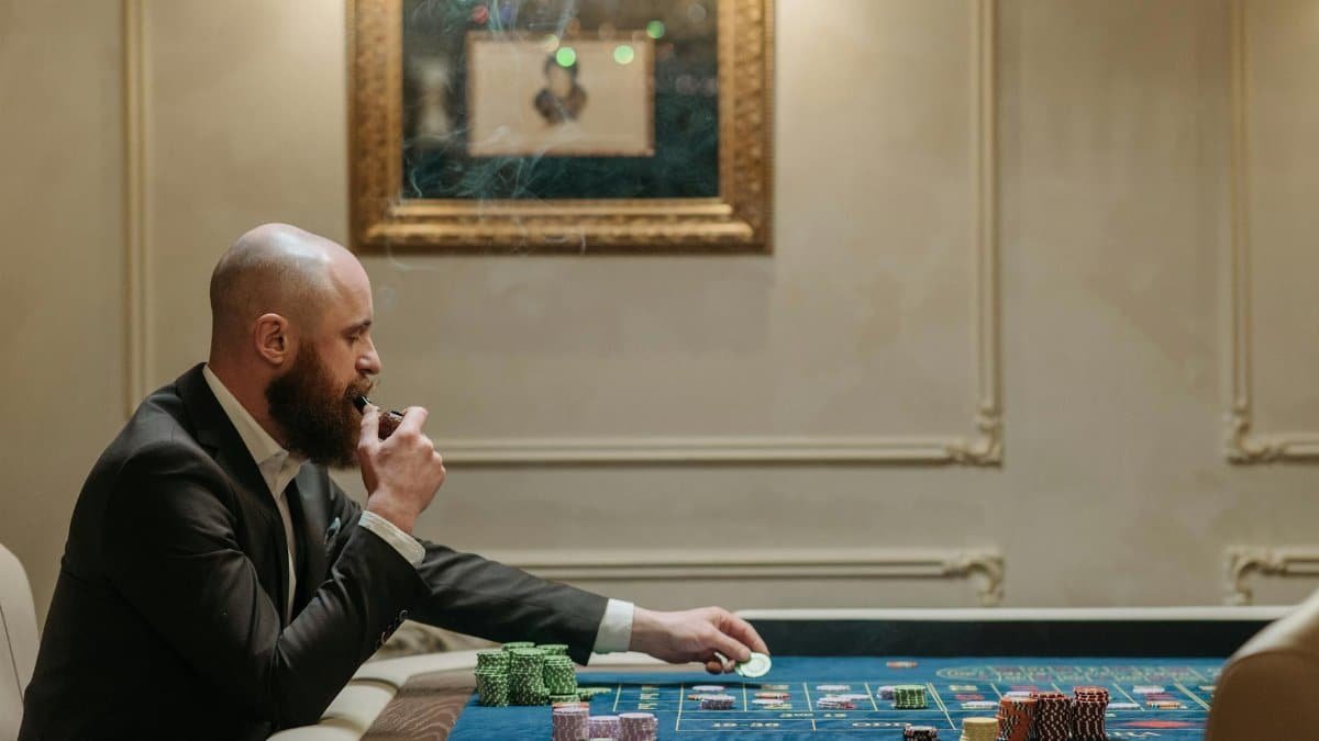 A man in a suit smokes a pipe while playing with poker chips at a casino table indoors.