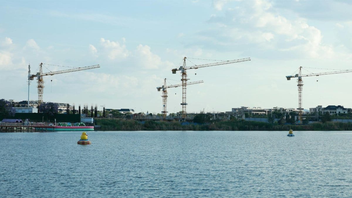 Construction cranes by a riverside under a clear sky, showcasing urban development.