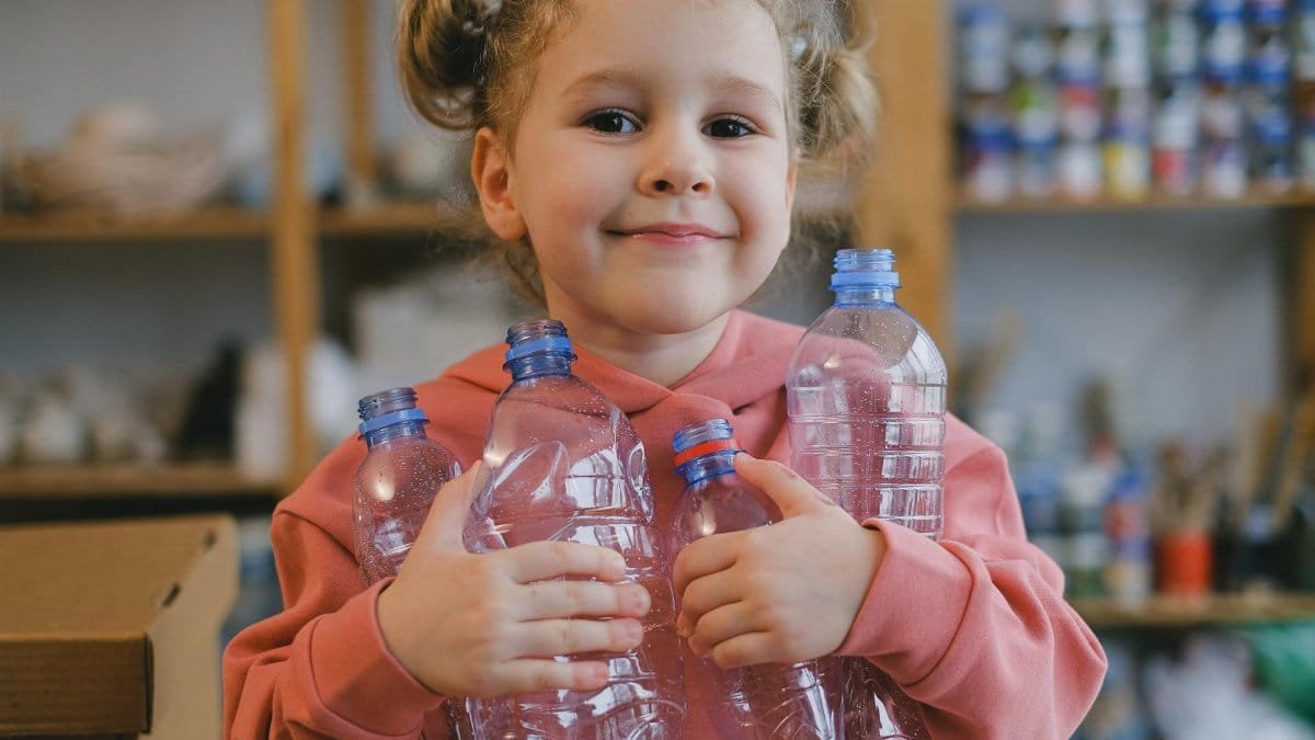 Happy child holding plastic bottles, smiling indoors, promoting recycling awareness.