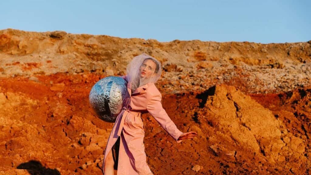 Woman in pink coat holding foil ball in vibrant desert, blending art and fashion.