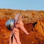 Woman in pink coat holding foil ball in vibrant desert, blending art and fashion.