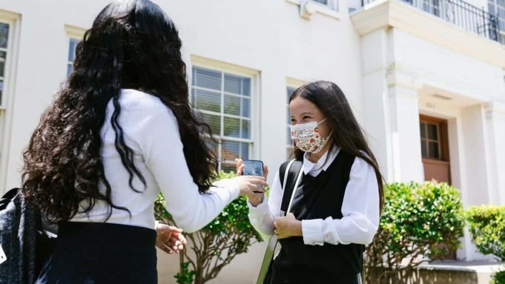 Two schoolgirls in uniforms wearing protective masks stand outside a school building.