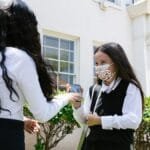 Two schoolgirls in uniforms wearing protective masks stand outside a school building.