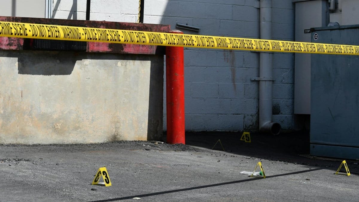 Sunlit urban scene showing a crime scene with caution tape and evidence markers, conveying investigation atmosphere.