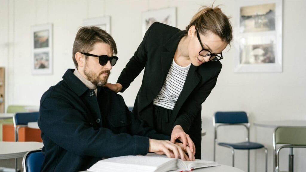 A visually impaired man reads Braille with the help of a caregiver in an indoor setting.