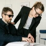 A visually impaired man reads Braille with the help of a caregiver in an indoor setting.