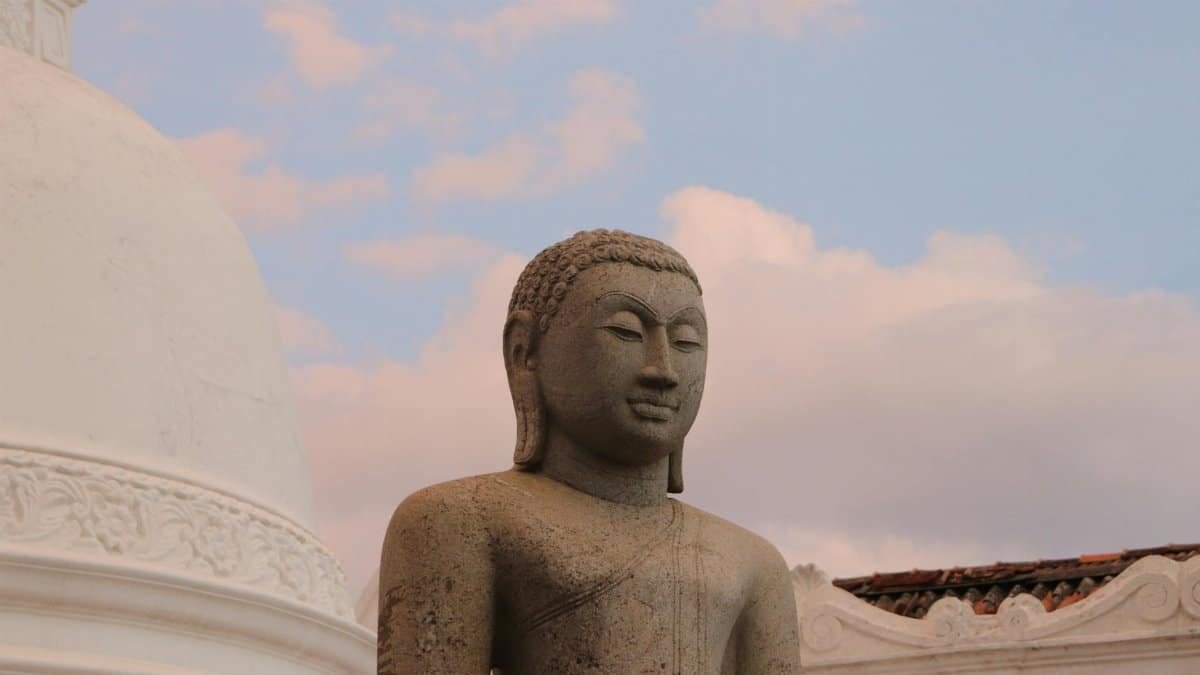 A tranquil stone Buddha statue against a vibrant sunrise sky near a white stupa.