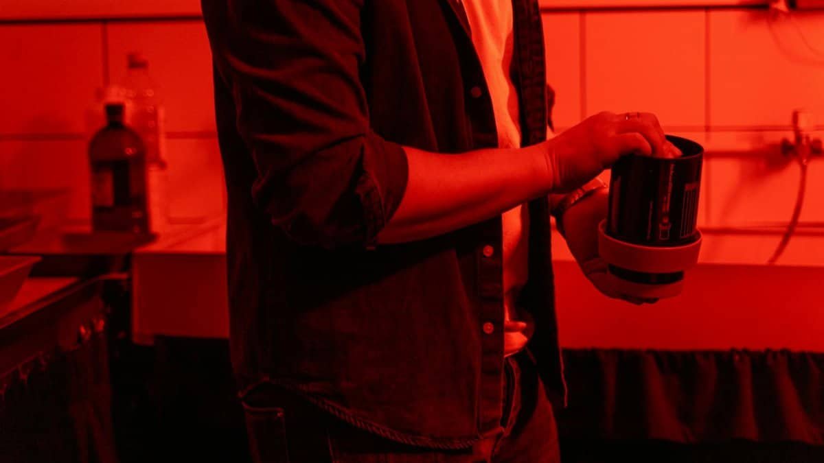 A photographer in a dimly lit darkroom processes film with chemical containers.