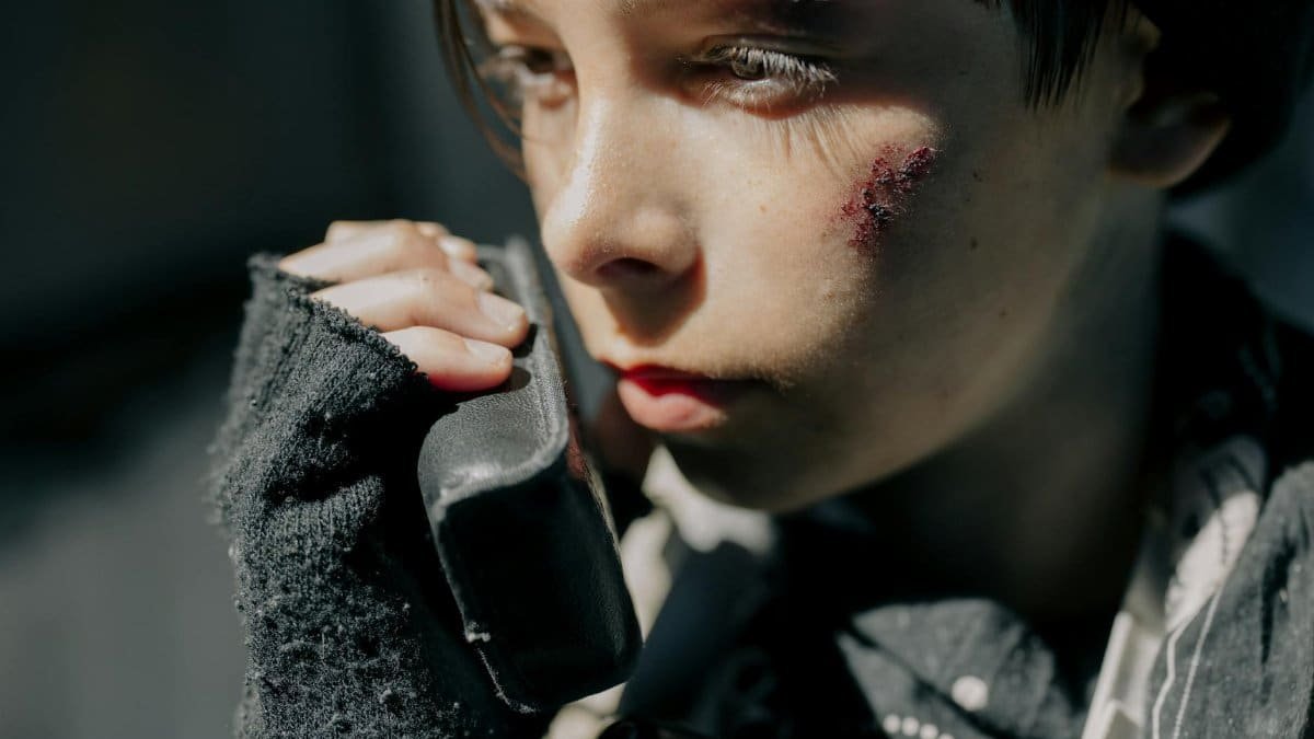 Close-up portrait of a young boy with wounds, using a radio, conveying a survival theme in a post-apocalyptic setting.