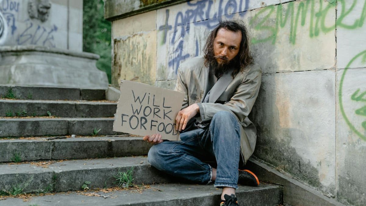 A homeless man sits on steps outdoors holding a 'Will Work for Food' sign, depicting poverty.