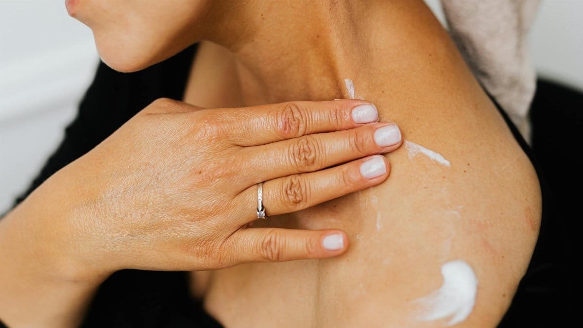 A close-up image of a woman applying skincare lotion on her neck with manicured hands.