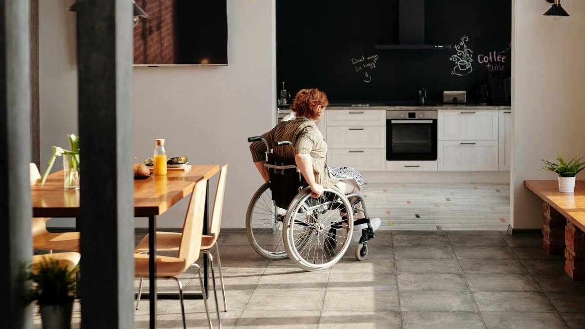 A woman in a wheelchair moves through a bright kitchen and dining area, emphasizing home accessibility.