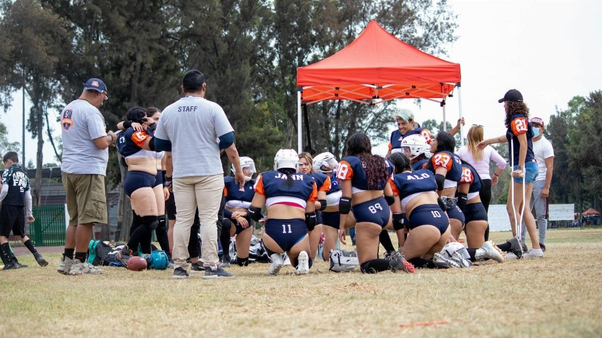 A women's flag football team huddles under a red canopy on a field, preparing for the game.