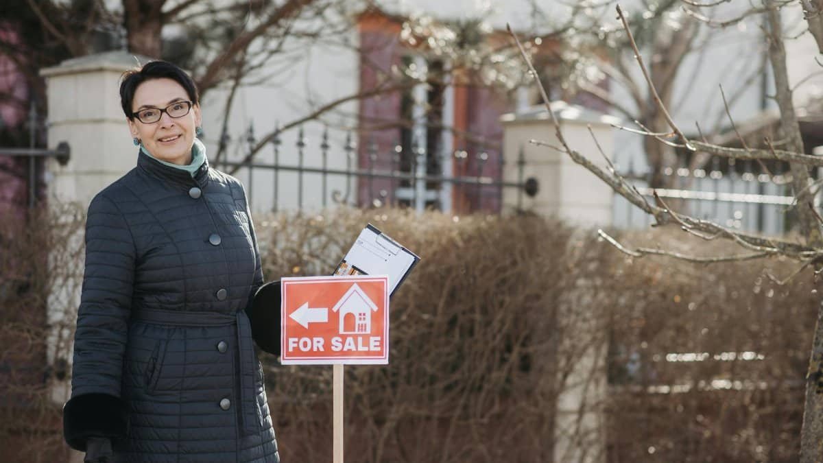Smiling woman realtor in winter coat standing by a for sale sign with documents, outdoors.