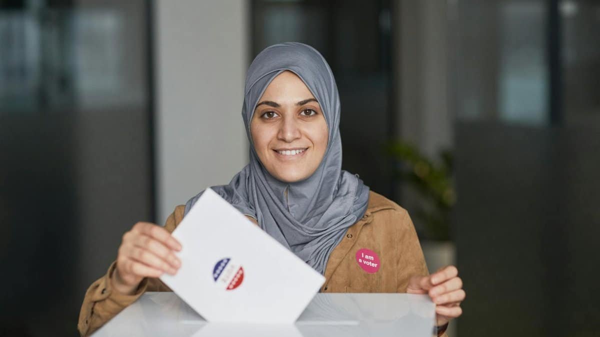 A smiling woman wearing a hijab casts her vote at a polling station indoors.