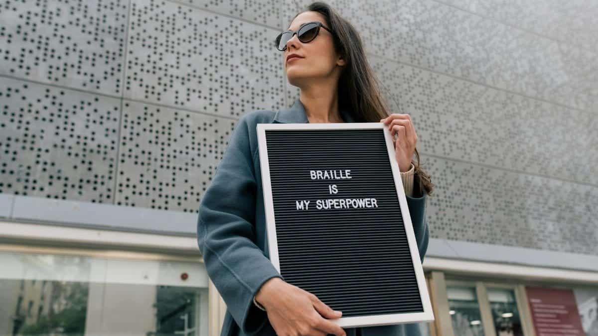 Woman in sunglasses holding a sign saying 'BRAILLE IS MY SUPERPOWER' outdoors.