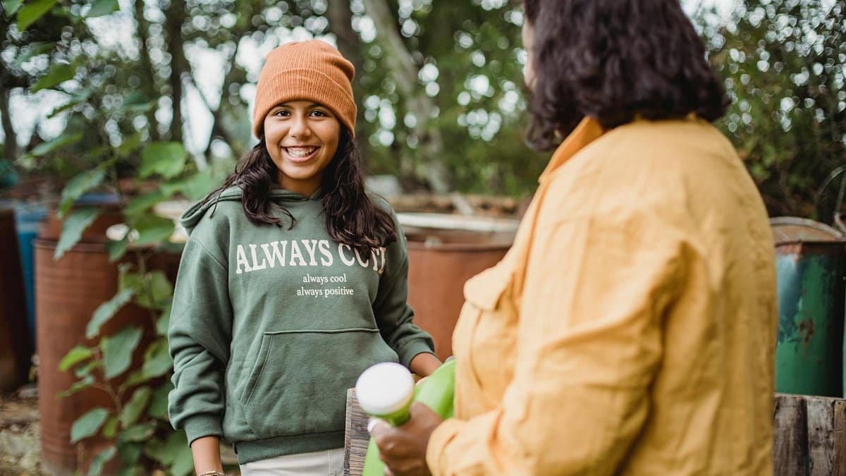 Happy ethnic teen looking at camera near crop anonymous farmer with watering can on farm on summer day