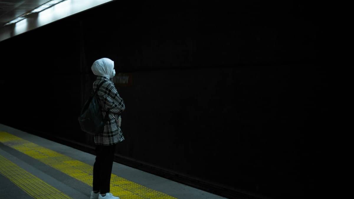 A woman in a hijab waits patiently at a dimly lit subway station platform at night.