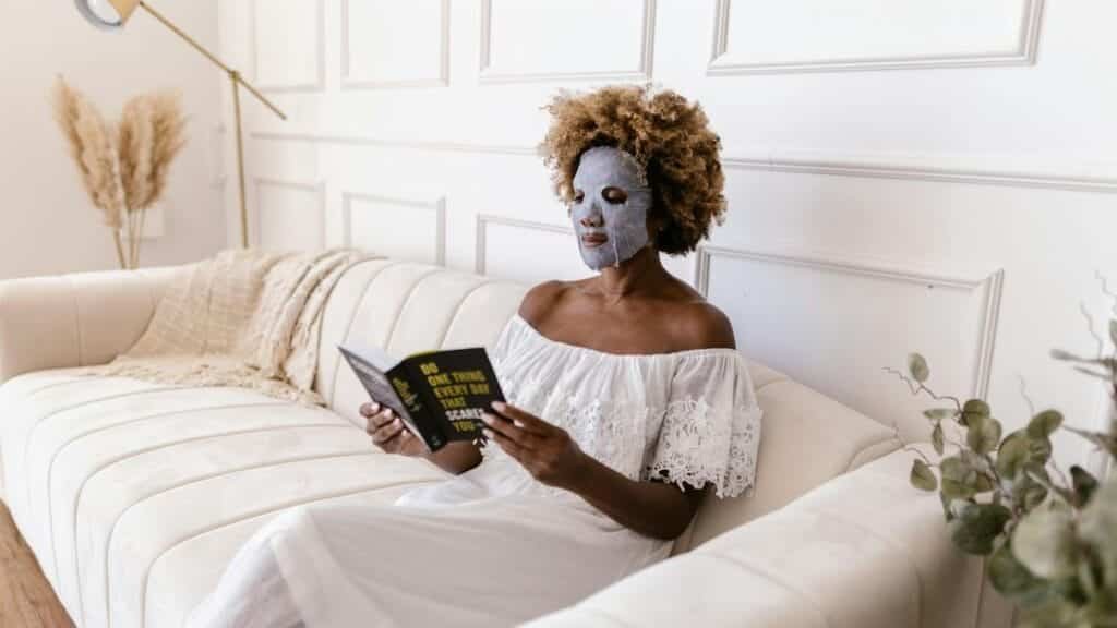 Woman with face mask reading a book on a cozy couch, embracing relaxation and self-care.