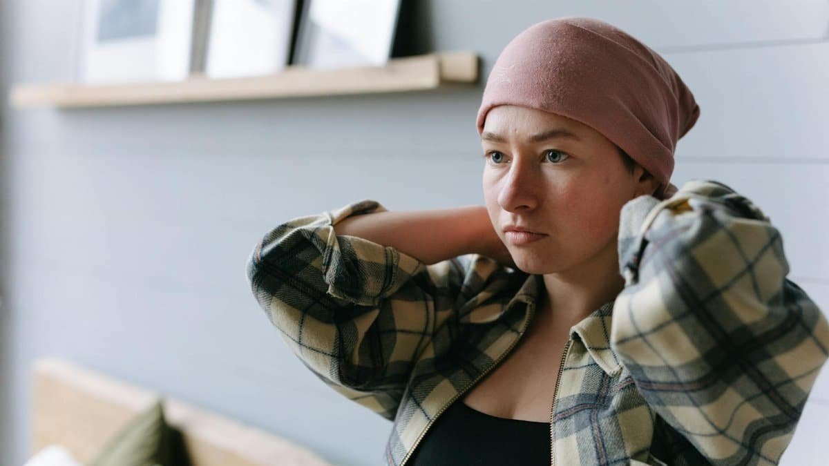 Woman with a pink headscarf adjusting her plaid shirt indoors, symbolizing strength and resilience.