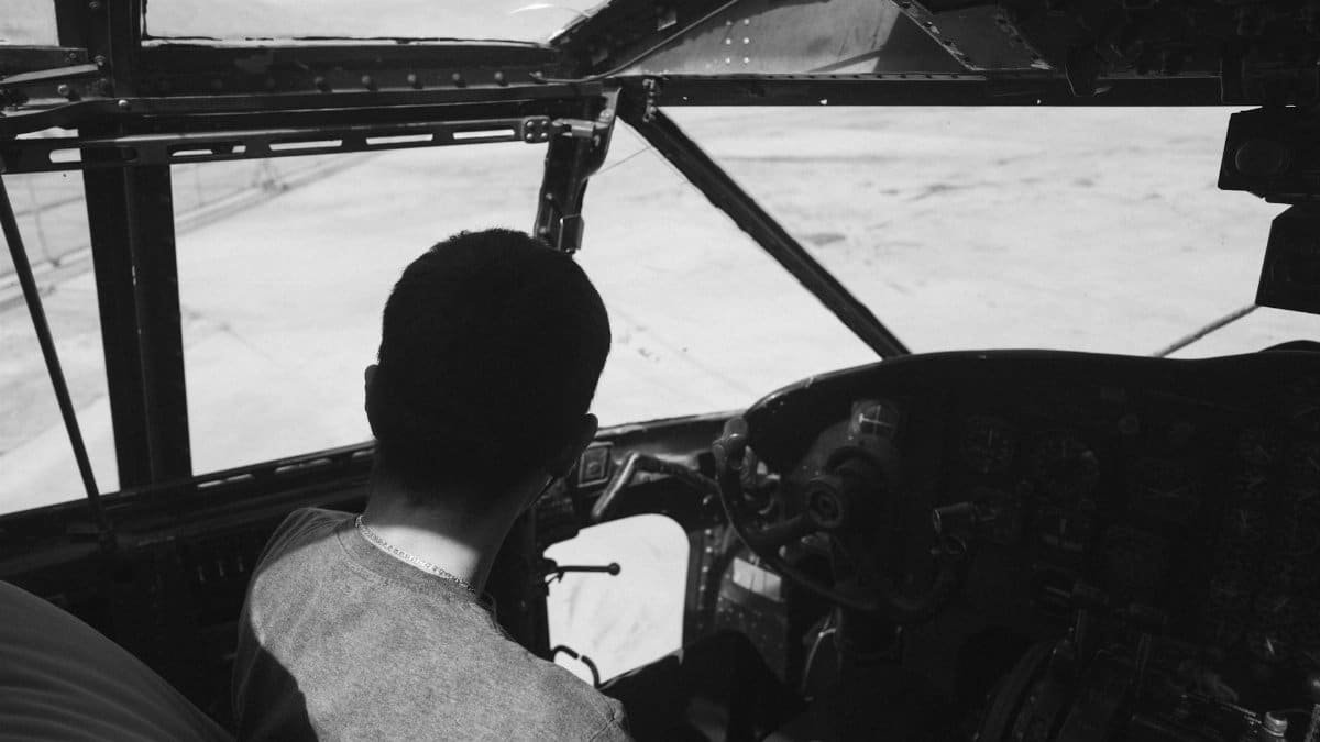 A black and white image showing a pilot navigating an aircraft cockpit from a rear view.