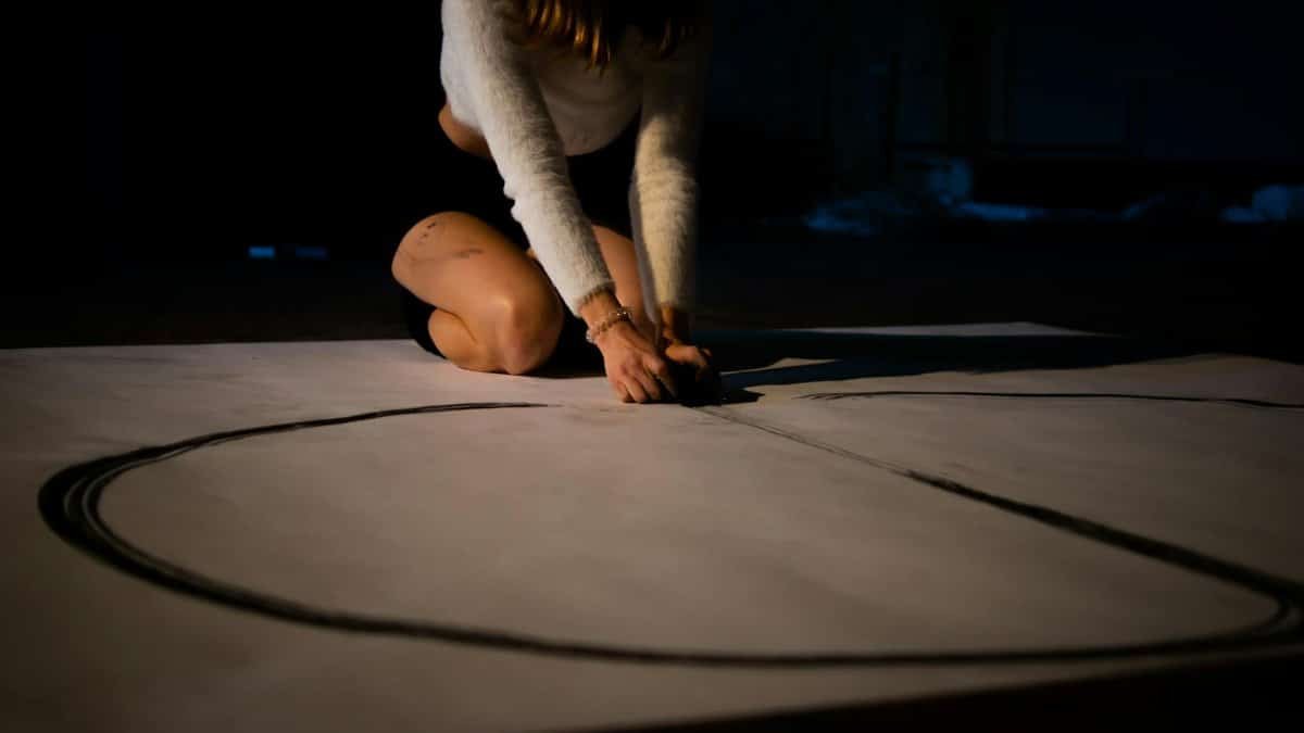 A woman drawing a large circle on the floor in a dimly lit space, focusing on artistic expression.