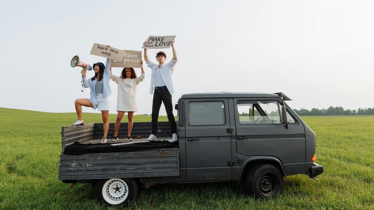 Three activists holding placards on a truck, advocating peace and love in a grassy field.