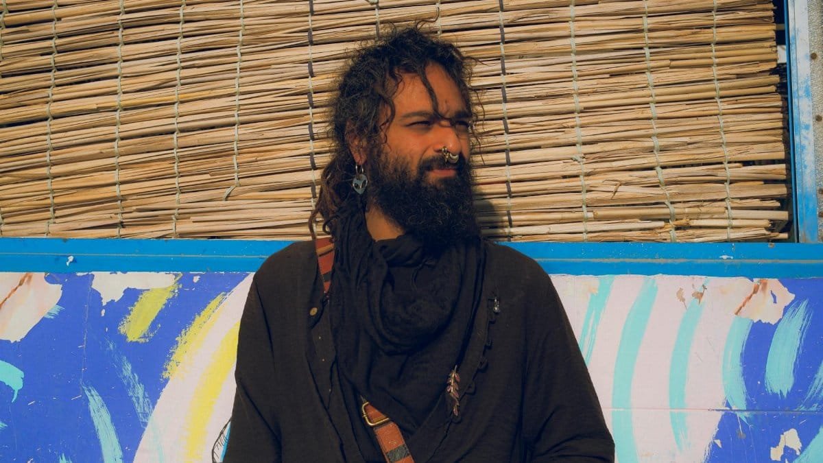 A bearded man standing against a colorful wall in bright daylight.