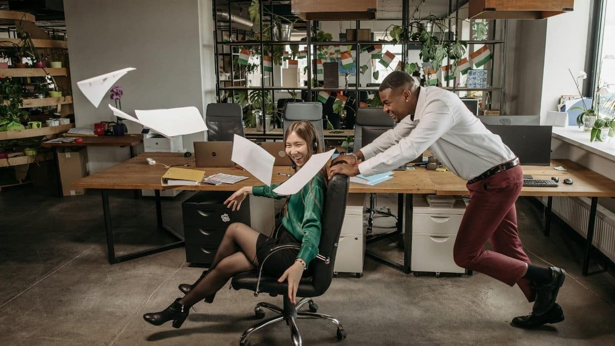 Two colleagues having fun in the office, pushing a chair amidst a lively work environment.
