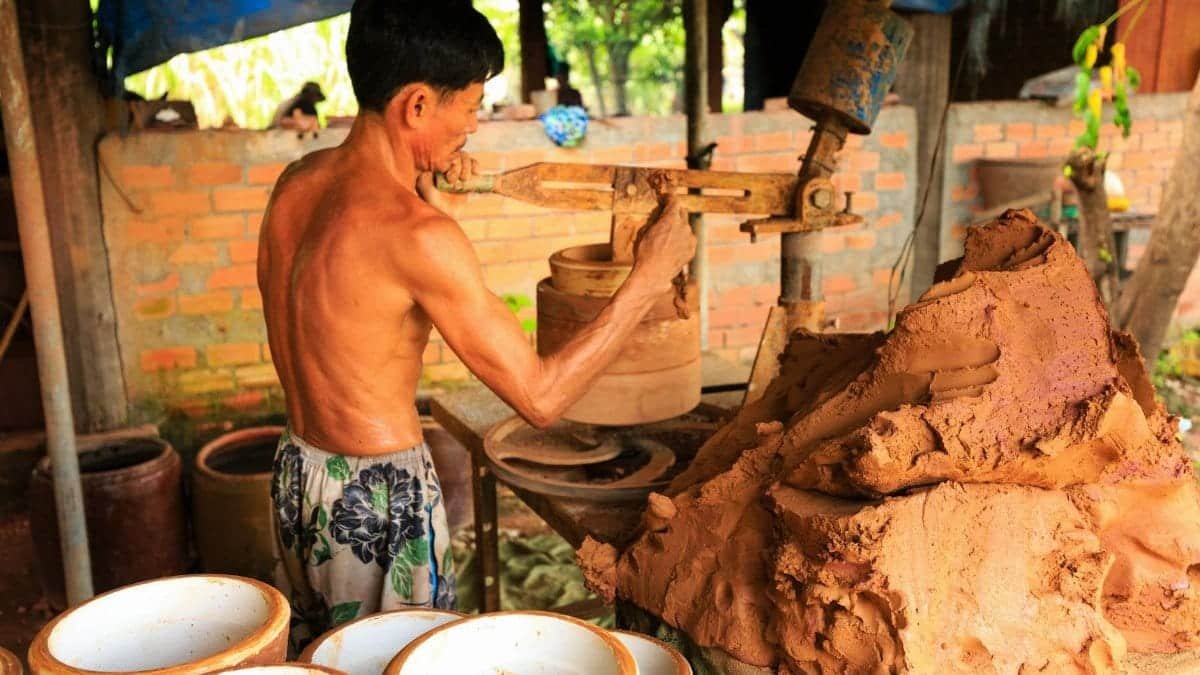 Shirtless Asian man skillfully crafting pottery in an outdoor setting with clay and earthenware.