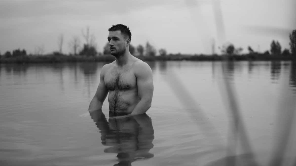 A contemplative black and white portrait of a shirtless man in a lake at dawn.
