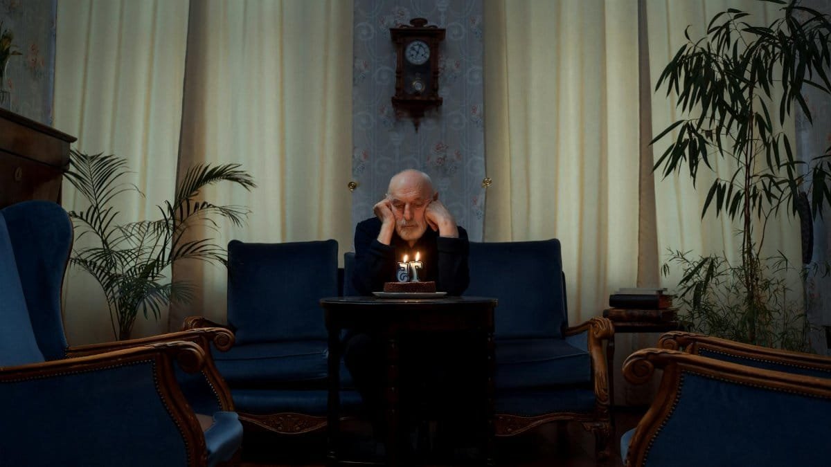 Senior man alone with a birthday cake in a vintage living room setting.