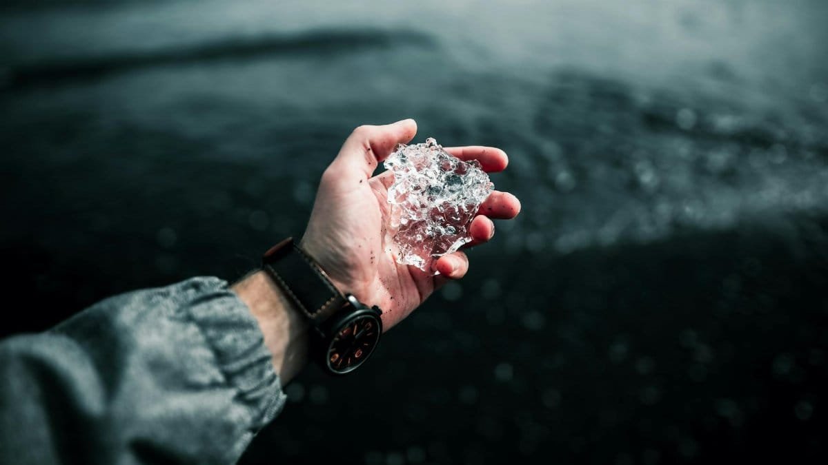 A hand with a wristwatch holding a piece of ice over dark sea water.